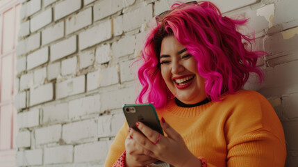 Fototapeta premium A person with vibrant pink hair dressed in a yellow sweater laughs while looking at their phone, leaning against a textured brick wall.
