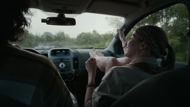 Medium rear shot of cheerful young Caucasian woman and man driving in car offroad during rain, on way to vacation, girl sitting with feet on dashboard, pointing to destination, chatting and smiling