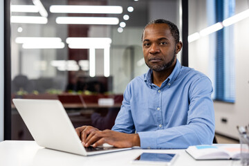 Obraz premium African American businessman sitting at desk with laptop inside modern office. Professional workplace setting with businessman focusing on work. Office setting promotes productivity professionalism.