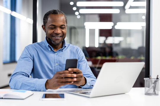 Mature african american office worker using smartphone modern workplace. Businessman interacting with mobile device, smiling during work. Technology facilitating professional communication in office