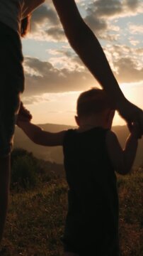 Vertical footage of caring mom helps her baby with first steps on background amazing mountains at sunset. Toddler child step concept. Mother helping son with his first steps in park. Selective focus