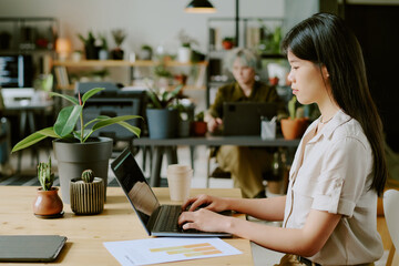 Side view of young Asian woman wearing casual clothes typing code on laptop at workplace in modern eco-friendly office with biophilic design