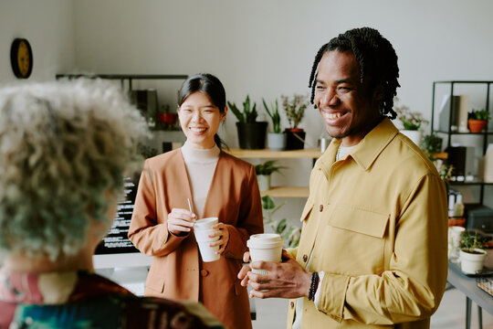 Group of three diverse gen Z colleagues standing in office during coffee break, laughing at funny joke