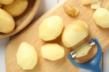 Fresh raw potatoes, peels and peeler on white wooden table, top view