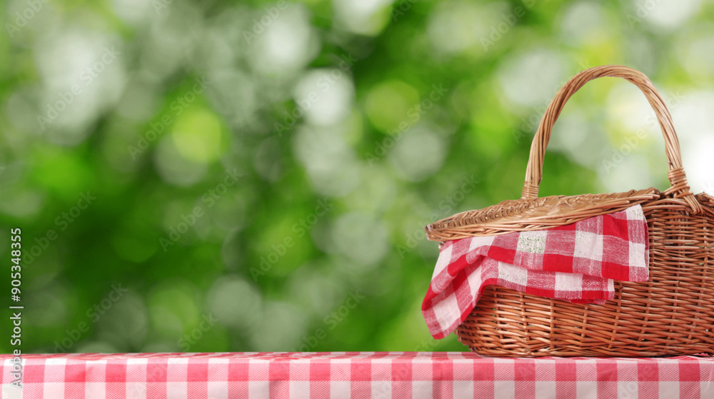 Wall mural Wicker picnic basket with red checkered napkin on table in nature, banner design. Space for text