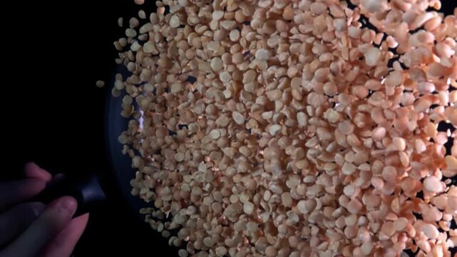 A hand holds the handle of a black frying pan with split peas flying out of the frying pan in super slow motion