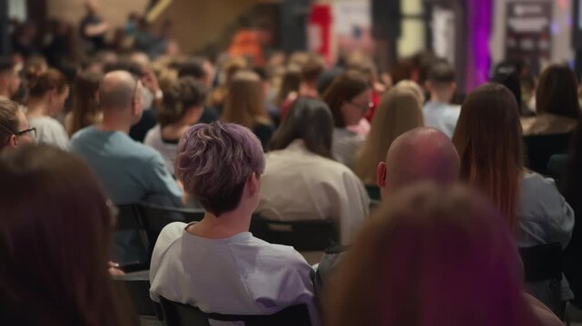 Girl with short colored haircut listens to report in a hall with a lot of people. Young professionals watch business incubator's pitch. Casual attire dominates this entrepreneurial assembly