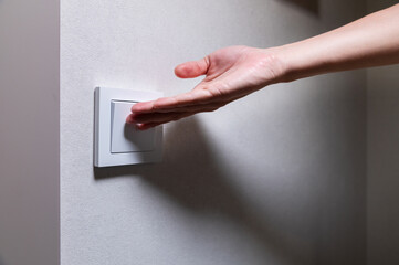 Close-up of an unrecognizable young Caucasian woman's hand turning on a light in a dark room by pressing a switch on a white wall. Close-up