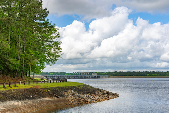 A wide-angle view of the West Point Lake Dame from Eagleview Park in Georgia against a backdrop of blue sky with large white clouds. Birds can be seen soaring in the sky.