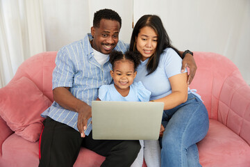 African American father and asian mother with daughter using laptop computer for watching movie or browsing internet on sofa