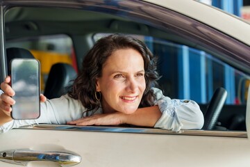 Caucasian woman adult sitting inside car leaning on window holding up smartphone with a smile. Showing positive interaction at repair shop, possibly related to car service or insurance.