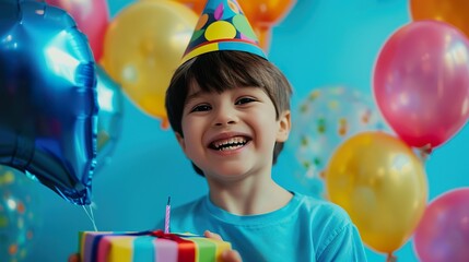 A happy boy in a blue birthday shirt, with a party hat and surrounded by colorful balloons, is eagerly holding a gift.