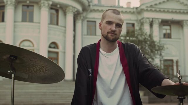 Zoom portrait shot of young male dummer posing for camera by drum set in park with historical building in background
