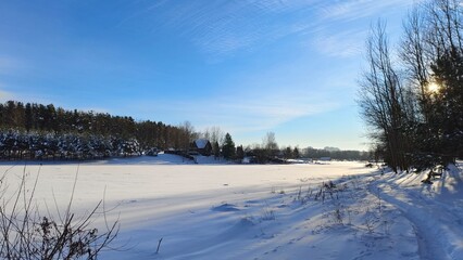 In winter, the lake is covered with ice and snow. There are trees on the shore, buildings on the far shore, and a path in the snowdrifts. The sun shines through the branches and the blue sky