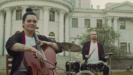 Zoom shot of young woman playing cello while man drumming in background during music performance in park in front of historical building