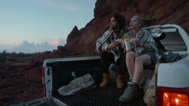 Full shot of cheerful young Caucasian man and woman sitting together in pickup trunk by red rock while traveling through desert, in twilight, snacking on sandwiches and tea, chatting and smiling
