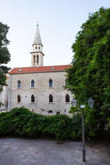 Naklejka premium An old stone church with a bell tower stands amidst lush green trees and a cobblestone courtyard adorned with street lamps.
