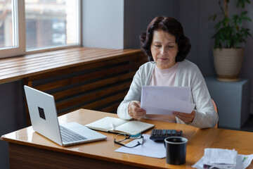 Middle aged senior woman sit with laptop and paper document, Pensive older mature lady reading paper bill pay online at home managing bank finances calculating taxes planning loan debt pension payment