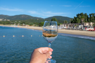 Hand with glass of cold rose wine from Provence and wooden yacht boota pier on white sandy beach Plage de Pampelonne near Saint-Tropez, summer vacation in France