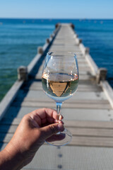 Hand with glass of cold rose wine from Provence and wooden yacht boota pier on white sandy beach Plage de Pampelonne near Saint-Tropez, summer vacation in France