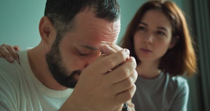 A sad and tired brunette guy with a beard leans on his hands and the girl tries to cheer him up and support him in the apartment