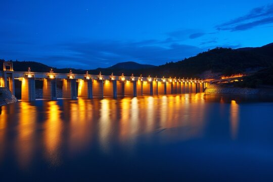 A long dam with multiple gates is illuminated at night, reflecting a warm glow on the calm water of the lake beneath. The sky is a deep blue with scattered stars.