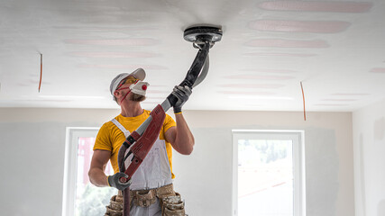 The worker sanding a plasterboard ceiling. He is using special drywall sander. A red plasterboard improve the fire resistance of ceiling structures.