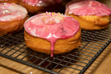 A selection of oil fried doughnuts topped with sweet pink and white icing and sprinkles on a baking trey