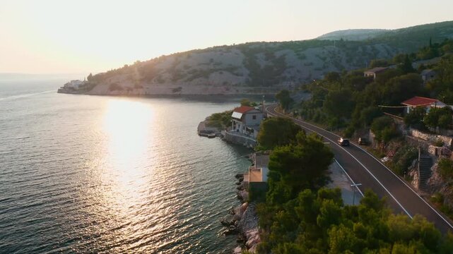 Passenger car driving alongside seashore. Sun reflects on sea bay water and road follows hill slope shape near mountains on shoreline aerial view