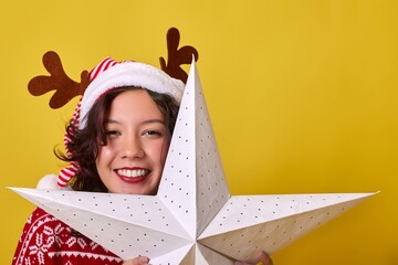 A young woman in a Santa hat with reindeer antlers holds a white star. She wears a cozy red and white striped sweater, radiating warmth and joy against a bright yellow background.