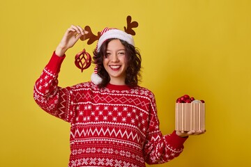 Young woman wearing a red sweater decorated with snowflakes and reindeer antlers is holding a red...