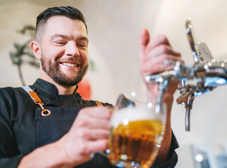 Smiling stylish bearded barman dressed black uniform with an apron tapping fresh lager beer into pilsner glass mug at bar counter. Successful people, beer consumption, beverages industry concept image