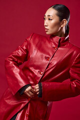 Fashionable asian woman in red leather coat posing with hands on hips against a red background