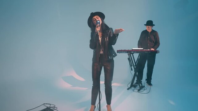 A vocalist in a black leather jacket and hat,smiling and singing with eyes open,stands at a vintage microphone with hands moving up and down A keyboardist works a red sampler against a blue background