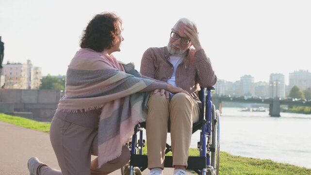 Old man in wheelchair walking with caregiver senior woman in park. Elderly family couple woman supporting taking care of paralyzed man in chair for people with disability outdoor. Rehabilitation