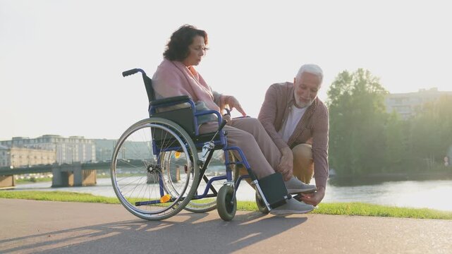 Senior woman in wheelchair walking with caregiver old man on road. Elderly family couple man supporting taking care of paralyzed woman in chair for people with disability outdoor. Rehabilitation