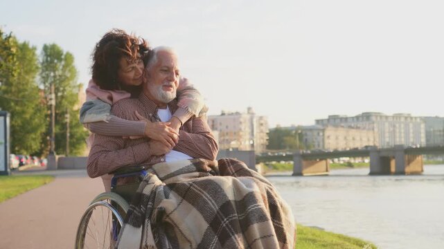 Old man in wheelchair walking with caregiver senior woman on road in park. Elderly family couple woman supporting embracing paralyzed man in chair for people with disability outdoor. Rehabilitation