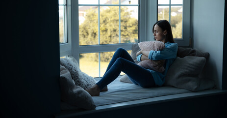 Sad, depressed, frustrated, angry, lonely, emotional young woman with severe stress and mental problem, disease, headache, illness, divorce, victim of violence. Female sitting on big window sill
