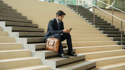 Smart project manager looking at mobile phone while sitting at stairs. Attractive caucasian business man working by using phone. Professional investor using phone to plan business strategy. Exultant.