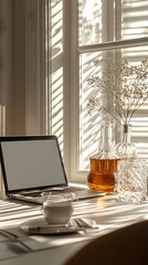 Front view home office workspace. Laptop computer with blank screen, glasses, decanter and glass of water on white table. Soft sunlight glare and shadows. Copy space