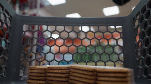 view of a lady in a black coat wearing a backpack placing a juice into a shopping trolley that already contains a pack of biscuits. Shot with a handheld camera