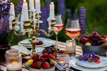 Elegant gorgeous wedding table decor or romantic dinner arrangement outdoors in blooming field. Purple lupine flowers, candles, fruits and wine, wooden vintage furniture. Sunset, summer, golden hour