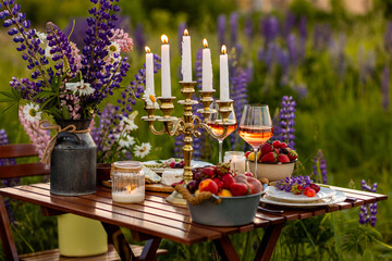 Simple countryside beautiful wedding decor, surprise romantic date outdoors, garden arrangement with lupins flowers on a meadow. Fruits, rose wine, burning candles. Sunset, candlelight, golden hour.