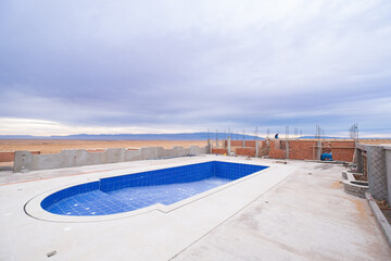 An empty swimming pool under construction in the Moroccan desert. It is dawning in the desert on a cloudy day. There are cement, bricks and construction materials