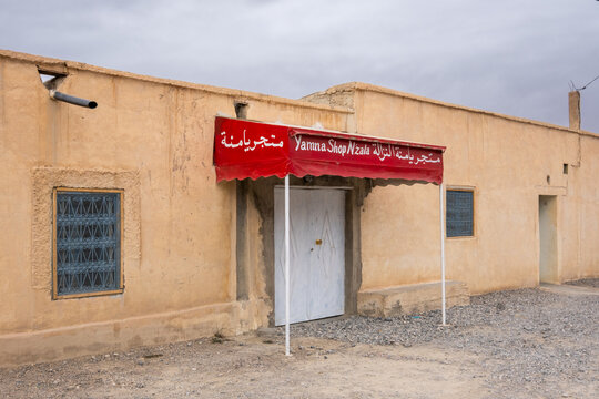 A small roadside bar with a red awning and a classic metal door typical of Morocco. It's in the Moroccan desert and it's a cloudy day.