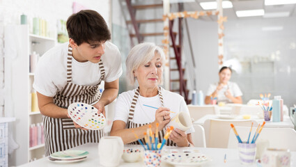 Positive young guy assisting enthusiastic senior woman painting ceramic mug in pottery class,...