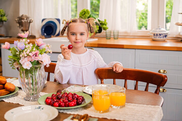 A charming girl eats ripe red cherries in the kitchen of a country house