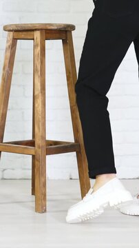 Model in black jeans demonstrating white massive shoes. Lady turns around to show the fashionable footwear. Wooden tabouret at backdrop. Vertical video