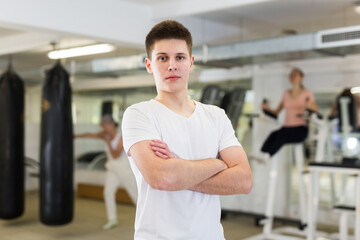 Confident athletic young guy posing in gym during workout, standing with crossed arms. Concept of promoting healthy active lifestyle among youth