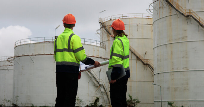 Male and female energy engineers collaborate with a laptop, blueprint, and digital tablet at the oil storage tanks in chemical and refinery  plant